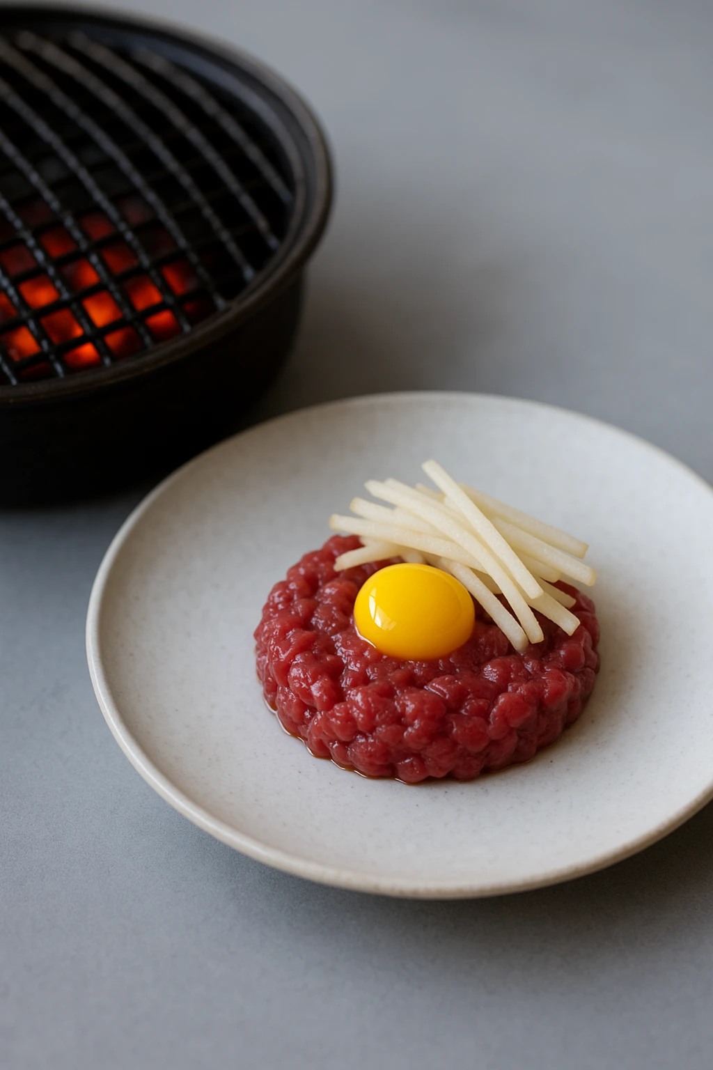 Beef tartare (yukhoe) with julienned pear and quail egg yolk presented next to hot grill for contrast — minimalist fine‑dining plating, cool neutral tones, large aperture.